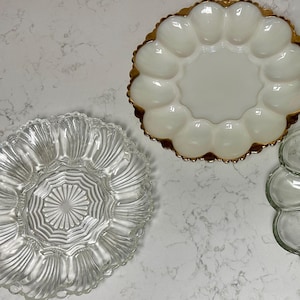 May include: Three round serving platters designed for eggs. One platter is made of opaque white milk glass with a scalloped gold-trimmed edge. The other two platters are made of clear pressed glass, featuring radial patterns and twelve indentations.