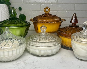 May include: A selection of glass candy dishes with lids in shades of green, amber, and clear. The dishes are filled with white wax and are displayed on a white surface. The lids feature decorative finials.