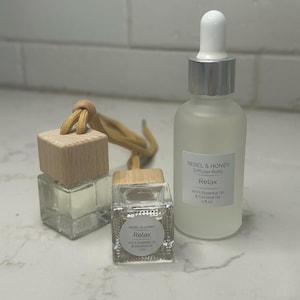 May include: Three glass bottles with wooden tops and the text "REBEL & HONEY Relax". One bottle is a diffuser refill with a dropper, and the others are car air fresheners. The bottles contain a clear liquid.