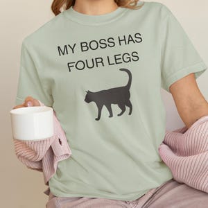 May include: A light green t-shirt with the text "MY BOSS HAS FOUR LEGS" in black. A black cat silhouette is printed below the text. The shirt is made of a soft material.