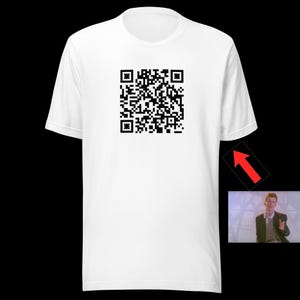 May include: White t-shirt with a large black QR code printed on the front. The shirt has short sleeves and a crew neck. A small image of a man in a suit is in the lower right corner, with a red arrow pointing to it.