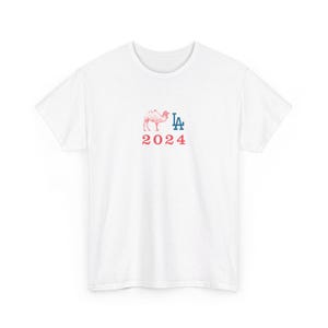 May include: White t-shirt with a red camel graphic and the blue letters "LA" above the red number "2024". The shirt is made of a soft material.