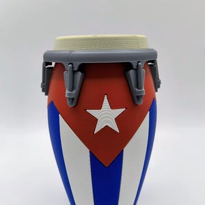 May include: A 3D-printed conga drum featuring the Cuban flag design. The drum has a red top section with a white star, blue and white stripes, and a gray rim and hardware. The drum is a musical instrument.