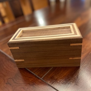 May include: A wooden box with a walnut finish and a light wood inlay. The box has a simple design with a rectangular shape and a lid that fits snugly.
