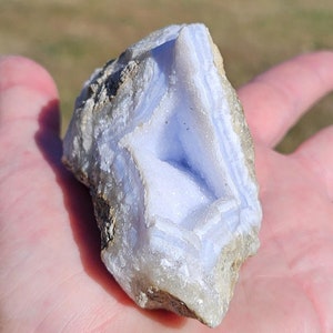 Puede incluir: Un trozo de piedra preciosa de &aacute;gata de encaje azul, sostenido en una mano. La piedra tiene un color azul claro con inclusiones blancas y beige. La superficie de la piedra tiene un aspecto en capas y bandeado, con una textura natural rugosa.