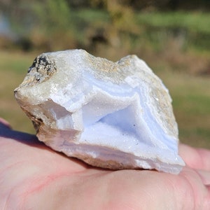 Puede incluir: Un trozo de &aacute;gata de encaje azul en bruto, una piedra semipreciosa, sostenido en una mano. La piedra muestra bandas de azul p&aacute;lido y blanco, con un exterior rugoso y texturizado. El fondo est&aacute; borroso, sugiriendo un entorno al aire libre.