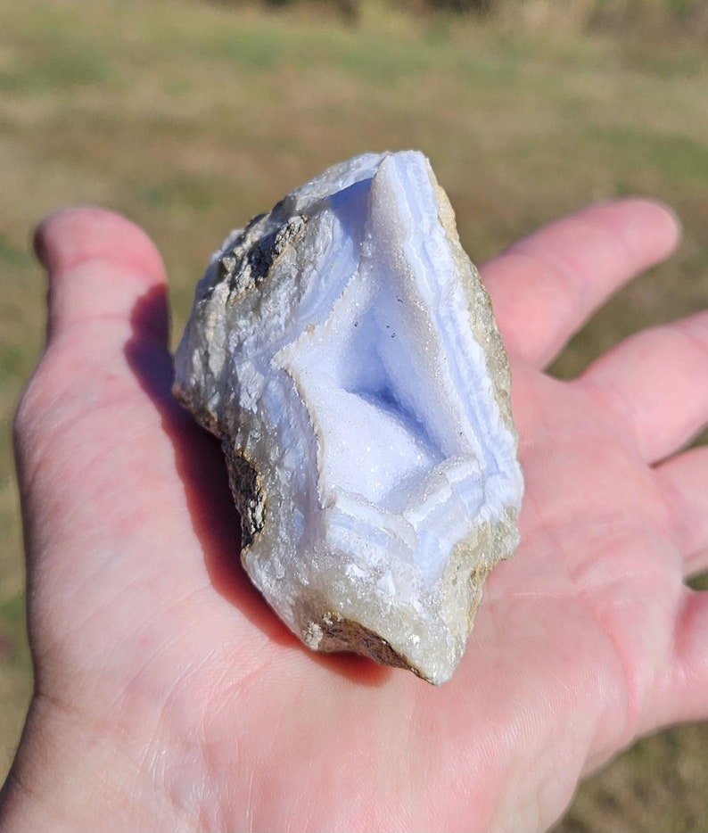 Puede incluir: Un trozo de piedra preciosa de &aacute;gata de encaje azul, sostenido en una mano. La piedra tiene un color azul claro con inclusiones blancas y beige. La superficie de la piedra tiene un aspecto en capas y bandeado, con una textura natural rugosa.