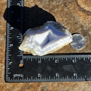 Puede incluir: Un esp&eacute;cimen de &aacute;gata de encaje azul con una moneda para comparar el tama&ntilde;o. La piedra tiene una apariencia en capas y bandeada con tonos de azul y blanco. La &aacute;gata mide aproximadamente 7,6 cm de largo.