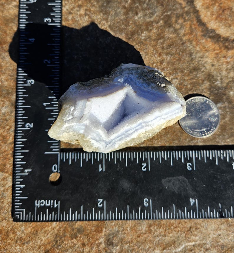 Puede incluir: Un esp&eacute;cimen de &aacute;gata de encaje azul con una moneda para comparar el tama&ntilde;o. La piedra tiene una apariencia en capas y bandeada con tonos de azul y blanco. La &aacute;gata mide aproximadamente 7,6 cm de largo.