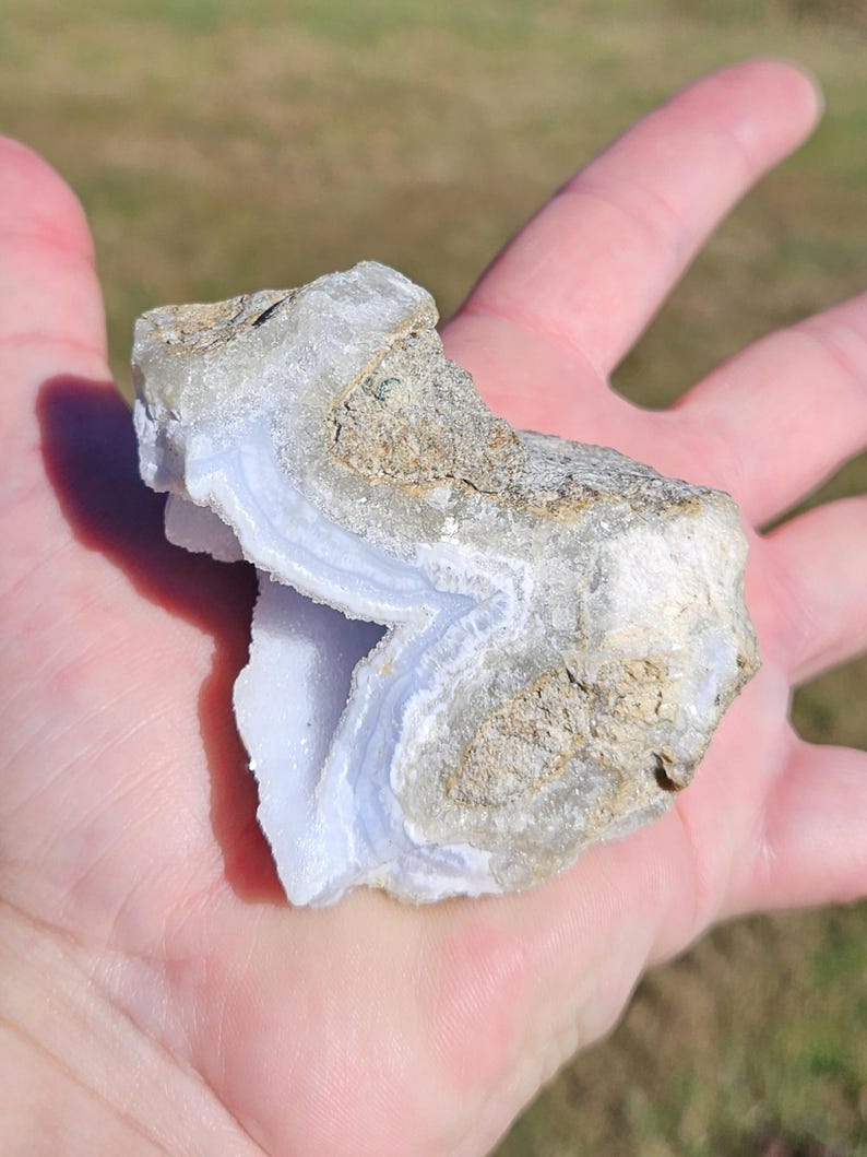Puede incluir: Un trozo de piedra preciosa de &aacute;gata de encaje azul, sostenido en una mano. La piedra tiene capas de azul p&aacute;lido y blanco, con un exterior rugoso y texturizado en tonos de beige y gris. La piedra preciosa tiene una forma natural y org&aacute;nica.