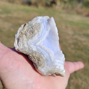 Puede incluir: Un trozo de &aacute;gata de encaje azul en bruto, una piedra semipreciosa, se sostiene en una mano. La piedra presenta bandas de azul p&aacute;lido y blanco, con un exterior rugoso y texturizado. El fondo est&aacute; borroso, lo que sugiere un entorno al aire libre.