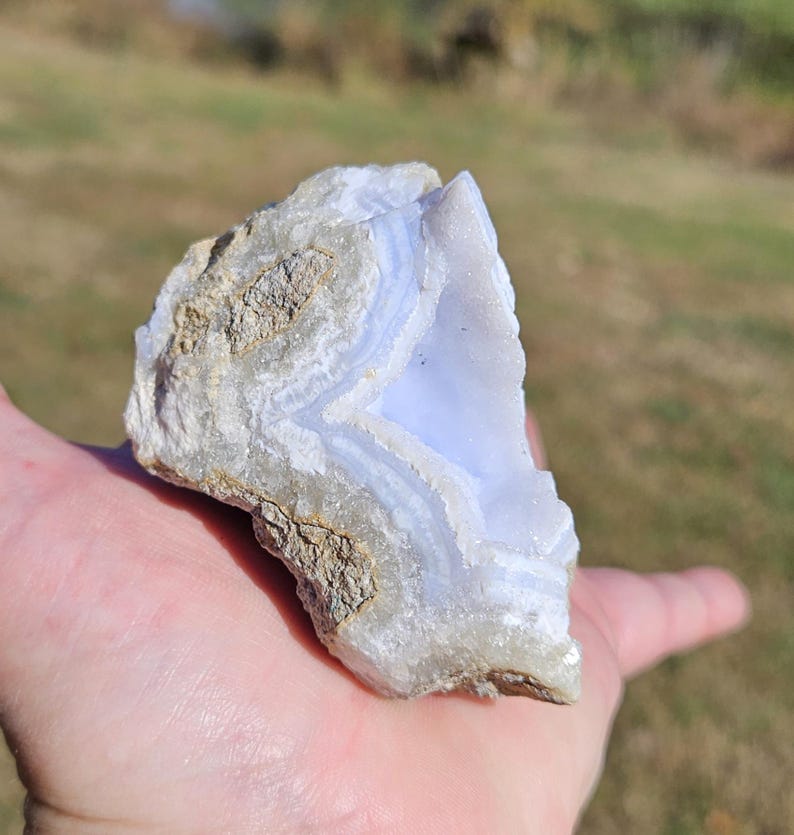 Puede incluir: Un trozo de &aacute;gata de encaje azul en bruto, una piedra semipreciosa, se sostiene en una mano. La piedra presenta bandas de azul p&aacute;lido y blanco, con un exterior rugoso y texturizado. El fondo est&aacute; borroso, lo que sugiere un entorno al aire libre.