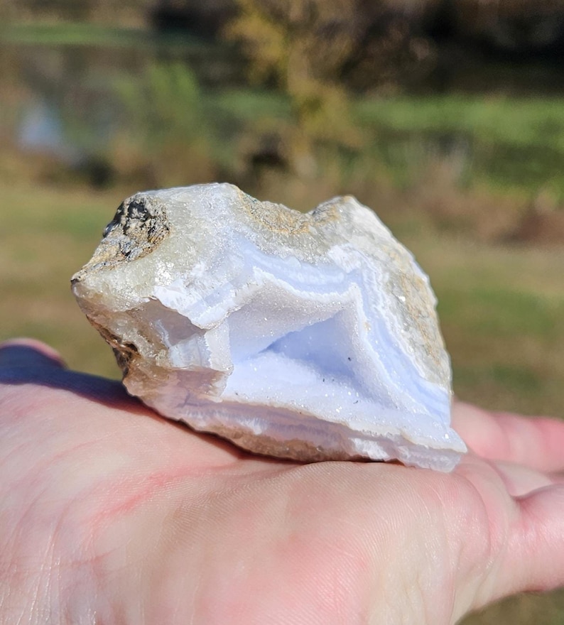 Puede incluir: Un trozo de &aacute;gata de encaje azul en bruto, una piedra semipreciosa, sostenido en una mano. La piedra muestra bandas de azul p&aacute;lido y blanco, con un exterior rugoso y texturizado. El fondo est&aacute; borroso, sugiriendo un entorno al aire libre.