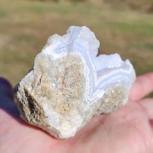 Puede incluir: Una piedra de &aacute;gata de encaje azul natural y en bruto, con un patr&oacute;n de bandas azul claro y blanco. La piedra se sostiene en una mano, mostrando su superficie rugosa y texturizada y sus formaciones cristalinas. La &aacute;gata tiene una mezcla de tonos beige y gris.