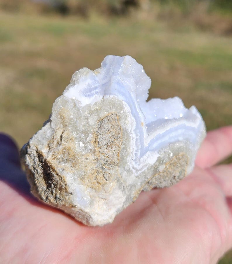 Puede incluir: Una piedra de &aacute;gata de encaje azul natural y en bruto, con un patr&oacute;n de bandas azul claro y blanco. La piedra se sostiene en una mano, mostrando su superficie rugosa y texturizada y sus formaciones cristalinas. La &aacute;gata tiene una mezcla de tonos beige y gris.
