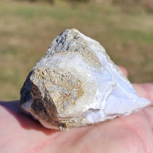 Puede incluir: Una gema de &aacute;gata de encaje azul natural, en bruto. La piedra muestra una mezcla de tonos azul claro, blanco y marr&oacute;n. El &aacute;gata tiene una estructura cristalina. Se sostiene en una mano, con un fondo borroso al aire libre.