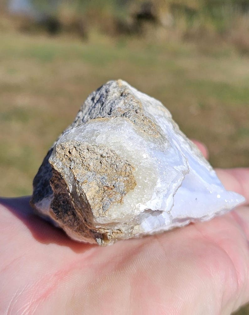 Puede incluir: Una gema de &aacute;gata de encaje azul natural, en bruto. La piedra muestra una mezcla de tonos azul claro, blanco y marr&oacute;n. El &aacute;gata tiene una estructura cristalina. Se sostiene en una mano, con un fondo borroso al aire libre.
