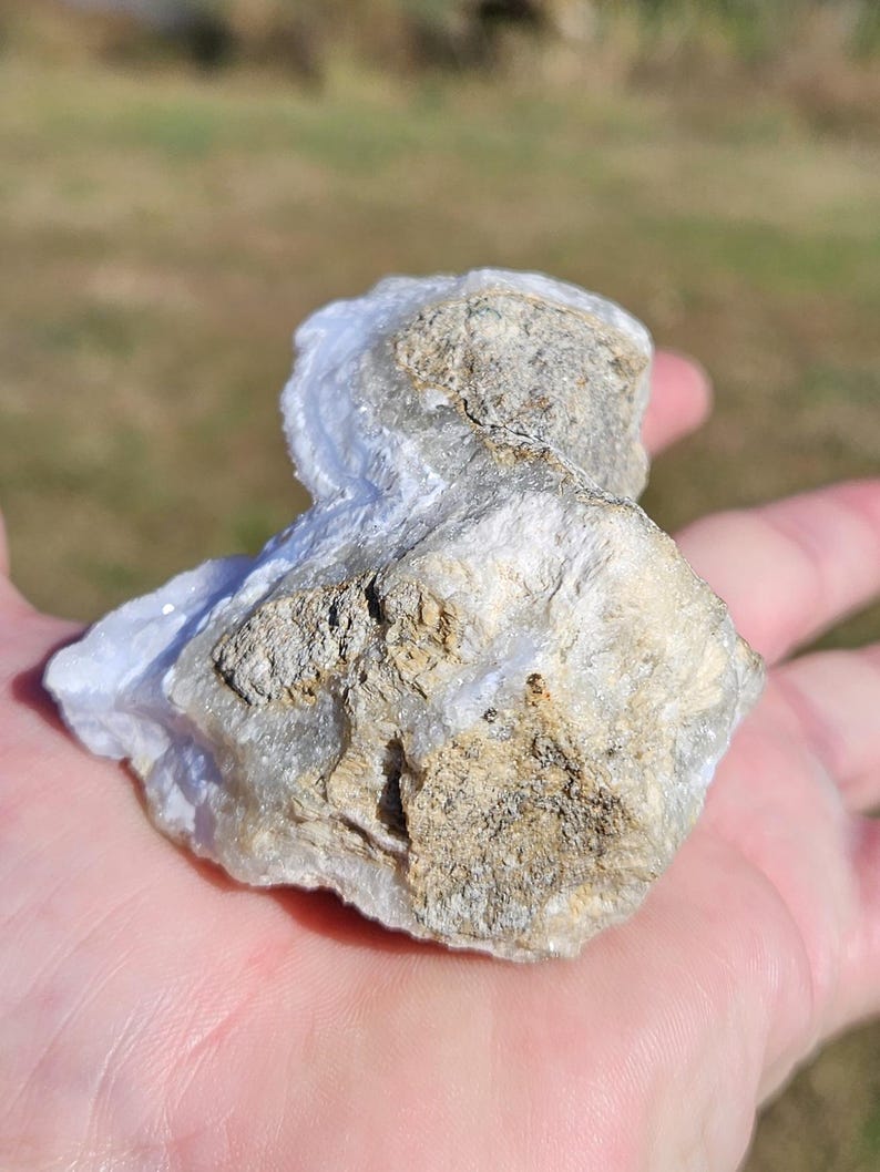 Puede incluir: Un trozo de &aacute;gata de encaje azul, una piedra preciosa natural, con una mezcla de colores azul claro y beige. La piedra tiene una superficie rugosa y texturizada y se sostiene en una mano.