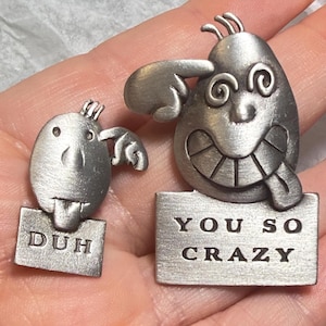 May include: Two silver-toned metal pins with a brushed finish. The smaller pin features a cartoon character with the text "DUH" on a rectangular base. The larger pin depicts a character with swirly eyes and a tongue out, with the text "YOU SO CRAZY".
