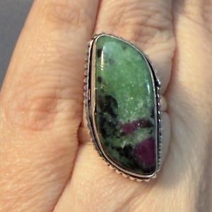 May include: A silver ring featuring a large, oval-shaped gemstone. The stone is primarily green with dark spots and patches of red. The ring has a detailed, textured border.