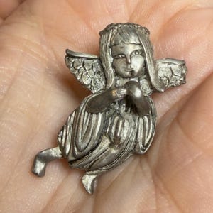 May include: A small, silver-colored angel figurine with detailed wings, a halo, and flowing robes. The angel is in a seated position, with hands clasped in prayer. The figurine is likely made of metal and has an antique or vintage appearance.