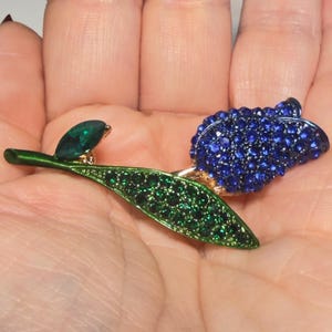 May include: A floral brooch featuring a blue flower with sparkling blue crystals, a green leaf with green crystals, and a green stem. The brooch has a gold-colored metal base and a green emerald-shaped crystal.