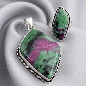 May include: A pendant and ring set featuring ruby zoisite gemstones. The pendant is a large, diamond-shaped stone with a silver rope-like border. The ring has a similar design, with a smaller, irregularly shaped stone. The gemstones display green, black, and pink hues.