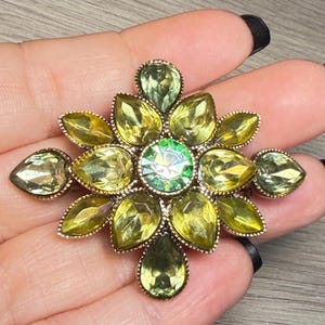 May include: A floral-shaped brooch featuring a central round green crystal surrounded by multiple pear-shaped yellow and olive green faceted crystals set in a metallic frame. The design radiates outward in a symmetrical pattern with pointed edges.