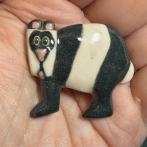 May include: A small ceramic panda figurine with a black and white striped body, black legs, and a white face with green eyes and a pink nose. The panda has small pink ears and is standing upright.