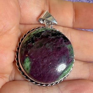 May include: A round pendant featuring a polished ruby and zoisite gemstone with deep purple and vibrant green inclusions. The stone is set in a silver-toned metal frame with a twisted rope-style border and a triangular bail at the top.