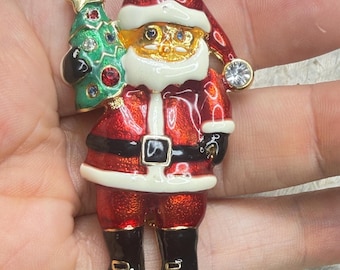 May include: A gold-tone metal brooch shaped like Santa Claus wearing a red suit with white trim and black boots. Santa holds a green Christmas tree decorated with small colorful rhinestones. A clear rhinestone accents the hat tip.
