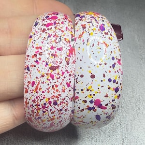 May include: Two thick, rounded bangle bracelets with a white base color, featuring a vibrant paint splatter pattern in shades of hot pink, bright yellow, deep purple, and dark red. The surface of the bangles is smooth and glossy with a reflective finish.