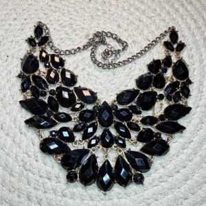 May include: A statement necklace featuring numerous faceted black teardrop and marquise-shaped stones set in a gold-tone metal frame. The design includes a central cluster of stones with a linked chain extending from the top sides.