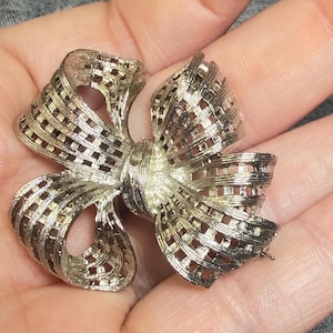 May include: A silver-toned brooch shaped like a bow with a textured, grid-like pattern throughout the ribbon loops. The metallic surface features a brushed finish with reflective highlights, creating a dimensional appearance across the entire bow design.