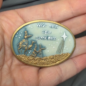 May include: An oval-shaped religious ornament with a gold border. The image depicts three wise men on camels, a star, and the text "wise men still seek him". The background is light blue with white stars.