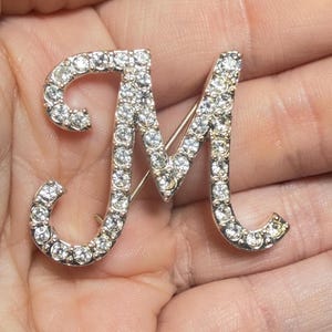 May include: A silver-toned metal brooch in the shape of the capital letter M, featuring a cursive script font. The entire surface of the letter is encrusted with numerous small, clear, round, faceted rhinestones that reflect light.