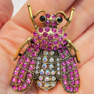 May include: A gold-toned metal bee brooch, adorned with vibrant pink and iridescent rhinestones. The insect-shaped pin features black eyes and detailed wings, creating a sparkling accessory.