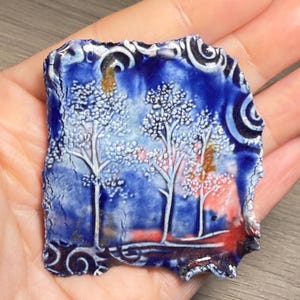 May include: A square ceramic tile featuring a hand-painted landscape of three white trees against a deep blue and red sky. The surface has a glossy, crackled glaze finish with decorative swirling patterns along the edges and a textured, uneven border.