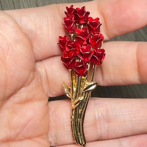 May include: A gold-tone metal brooch shaped like a bouquet of red enameled roses. The piece features a cluster of multiple deep red flower heads at the top, with long, smooth gold stems extending downward and two small gold leaves attached to the sides.