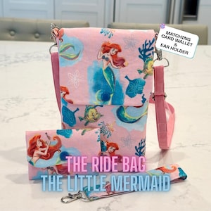 May include: A pink crossbody bag and matching wallet featuring a Little Mermaid print with Ariel and Flounder. The bag has a flap closure and an adjustable strap. Text on the bag reads: THE RIDE BAG THE LITTLE MERMAID. A tag reads: MATCHING CARD WALLET & EAR HOLDER.