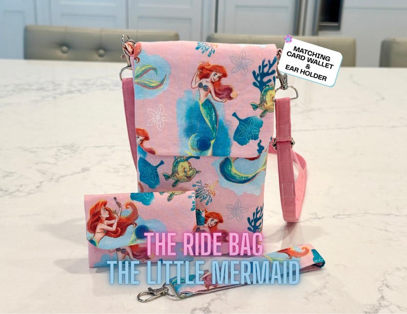 May include: A pink crossbody bag and matching wallet featuring a Little Mermaid print with Ariel and Flounder. The bag has a flap closure and an adjustable strap. Text on the bag reads: THE RIDE BAG THE LITTLE MERMAID. A tag reads: MATCHING CARD WALLET & EAR HOLDER.