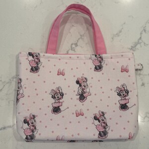 May include: A rectangular tote bag with pink handles and a zipper closure. The bag is covered in a pattern of Minnie Mouse illustrations and pink polka dots on a light pink background. The bag is designed for carrying items.