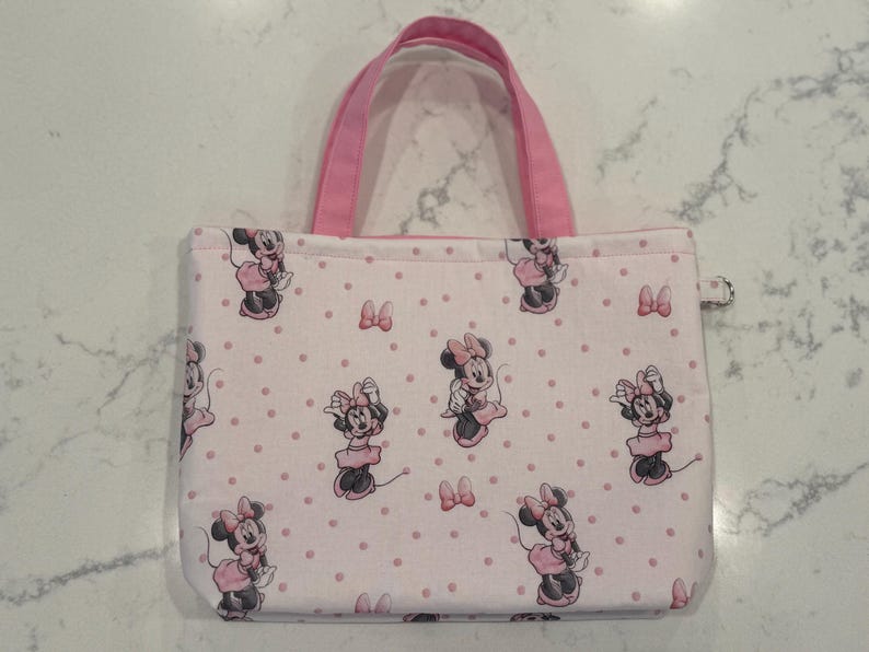May include: A rectangular tote bag with pink handles and a zipper closure. The bag is covered in a pattern of Minnie Mouse illustrations and pink polka dots on a light pink background. The bag is designed for carrying items.