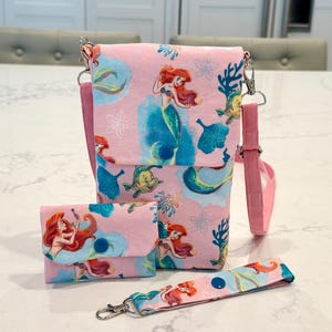 May include: A pink fabric crossbody bag, a matching small wallet with a blue snap closure, and a wristlet strap. The fabric features a repeating pattern of a red-haired mermaid, a yellow fish, blue coral, and white flowers on a light pink background.
