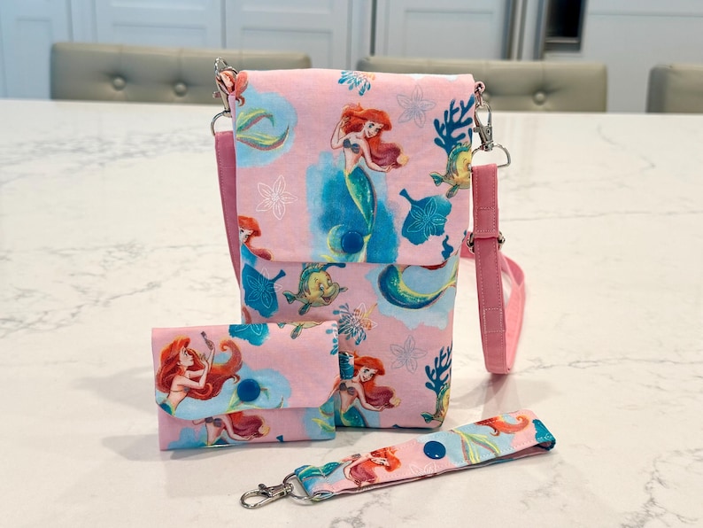 May include: A pink fabric crossbody bag, a matching small wallet with a blue snap closure, and a wristlet strap. The fabric features a repeating pattern of a red-haired mermaid, a yellow fish, blue coral, and white flowers on a light pink background.