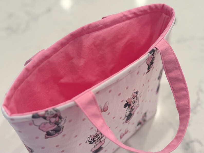 May include: Pink tote bag with a Minnie Mouse pattern and pink handles. The bag features a pink interior and a white exterior with pink polka dots and Minnie Mouse illustrations.
