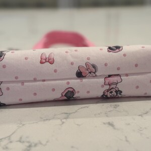 May include: A rectangular, pink fabric pencil case featuring a repeating pattern of Minnie Mouse illustrations and pink polka dots. The case has a matching pink handle and is likely used for storing school supplies or cosmetics.