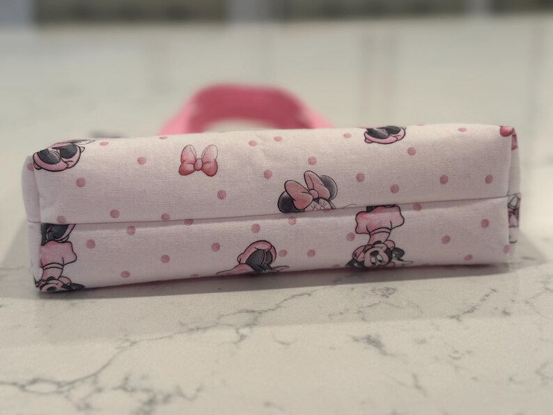 May include: A rectangular, pink fabric pencil case featuring a repeating pattern of Minnie Mouse illustrations and pink polka dots. The case has a matching pink handle and is likely used for storing school supplies or cosmetics.
