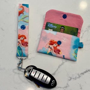 May include: A fabric wristlet keychain and a matching small wallet featuring a pink and blue pattern with an illustration of a mermaid. The wallet has a blue snap closure and contains a Disney Premier card. A black car key fob is attached to the wristlet.