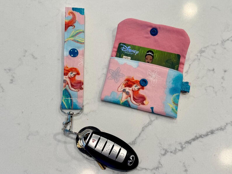 May include: A fabric wristlet keychain and a matching small wallet featuring a pink and blue pattern with an illustration of a mermaid. The wallet has a blue snap closure and contains a Disney Premier card. A black car key fob is attached to the wristlet.